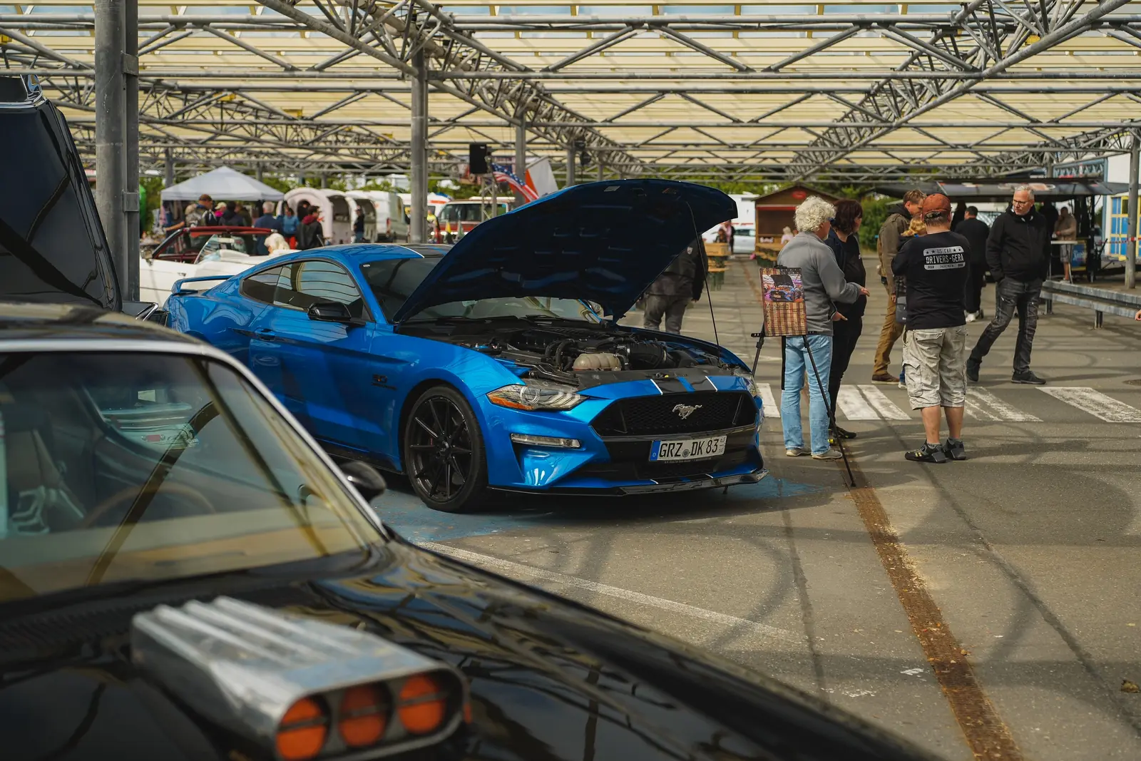 Blauer Mustang mit offener Haube unter dem Glasdach, Besucher schauen zu