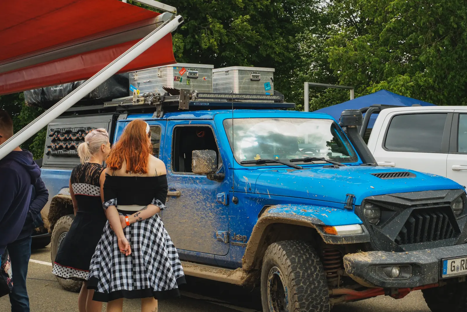 Zwei Besucherinnen im Fünfziger-Jahre-Kleid vor einem schlammigen blauen Jeep