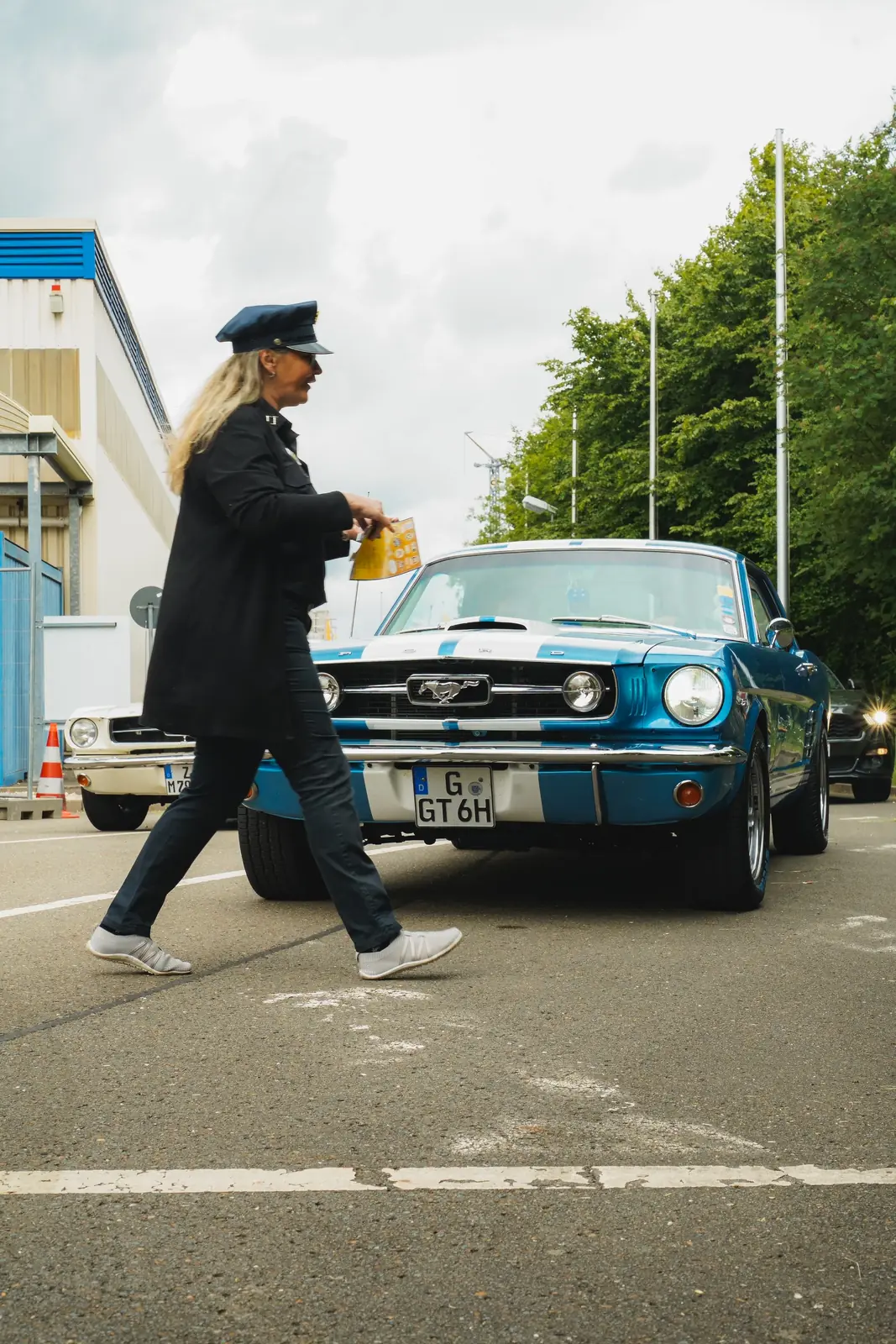 Frau in Uniform läuft vor einem blauen Mustang über die Zufahrt