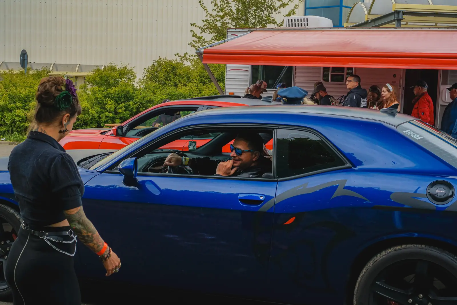 Fahrer im blauen Dodge Challenger lacht, davor eine Frau mit Tattoos