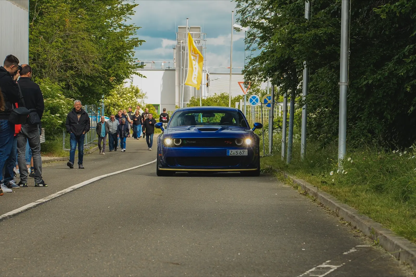 Blauer Dodge Challenger rollt die Zufahrt entlang, am Rand Besucher