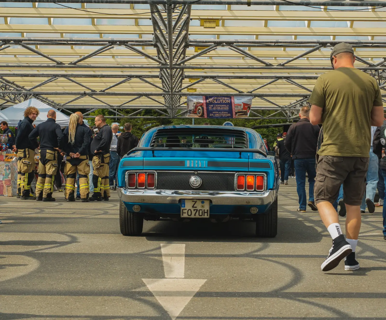 Blauer Mustang aus den Siebzigern von hinten, links Leute in Feuerwehrmontur