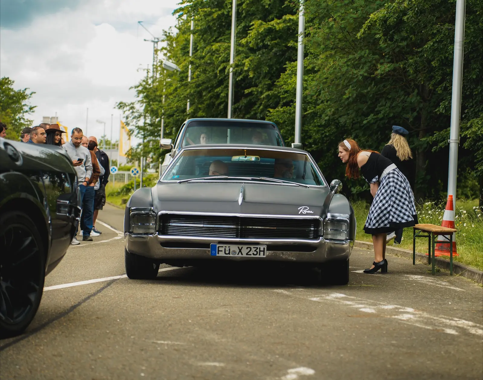 Dunkler Buick Riviera an der Einfahrt, daneben eine Besucherin im karierten Kleid