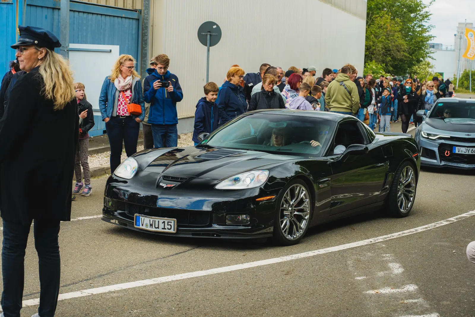 Schwarze Corvette fährt an einer Menschenmenge vorbei, vorn eine Frau in Uniform