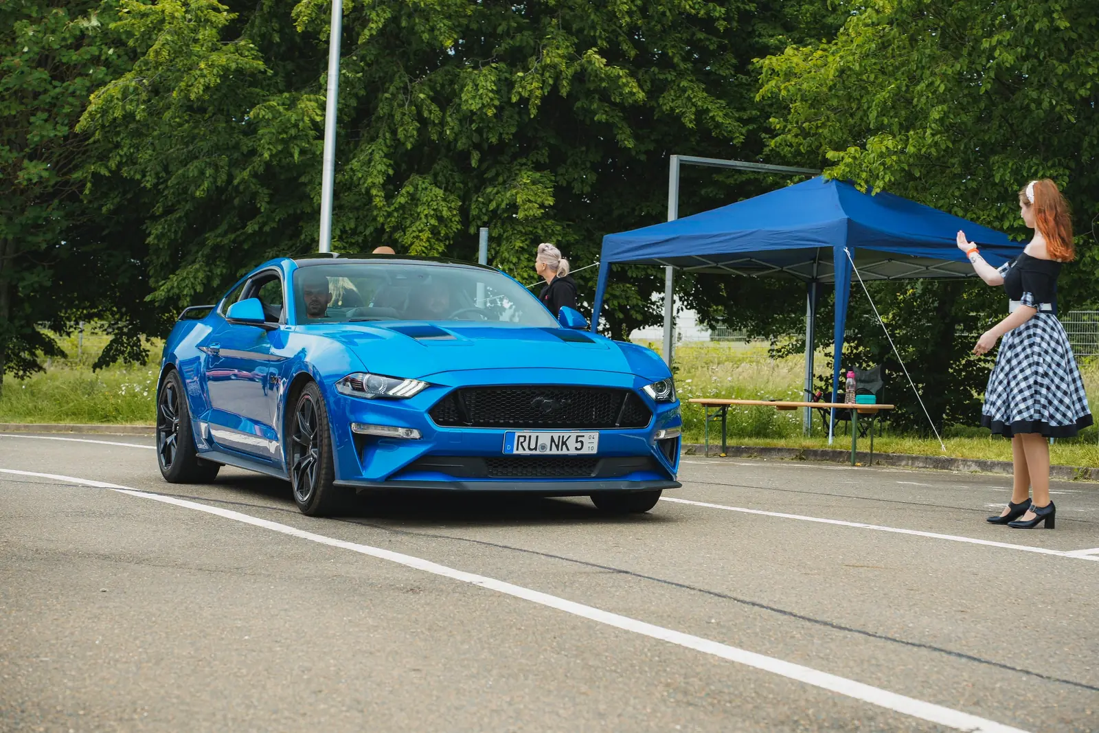 Blauer Mustang auf dem Platz, eine Frau im Petticoat winkt ihn ein