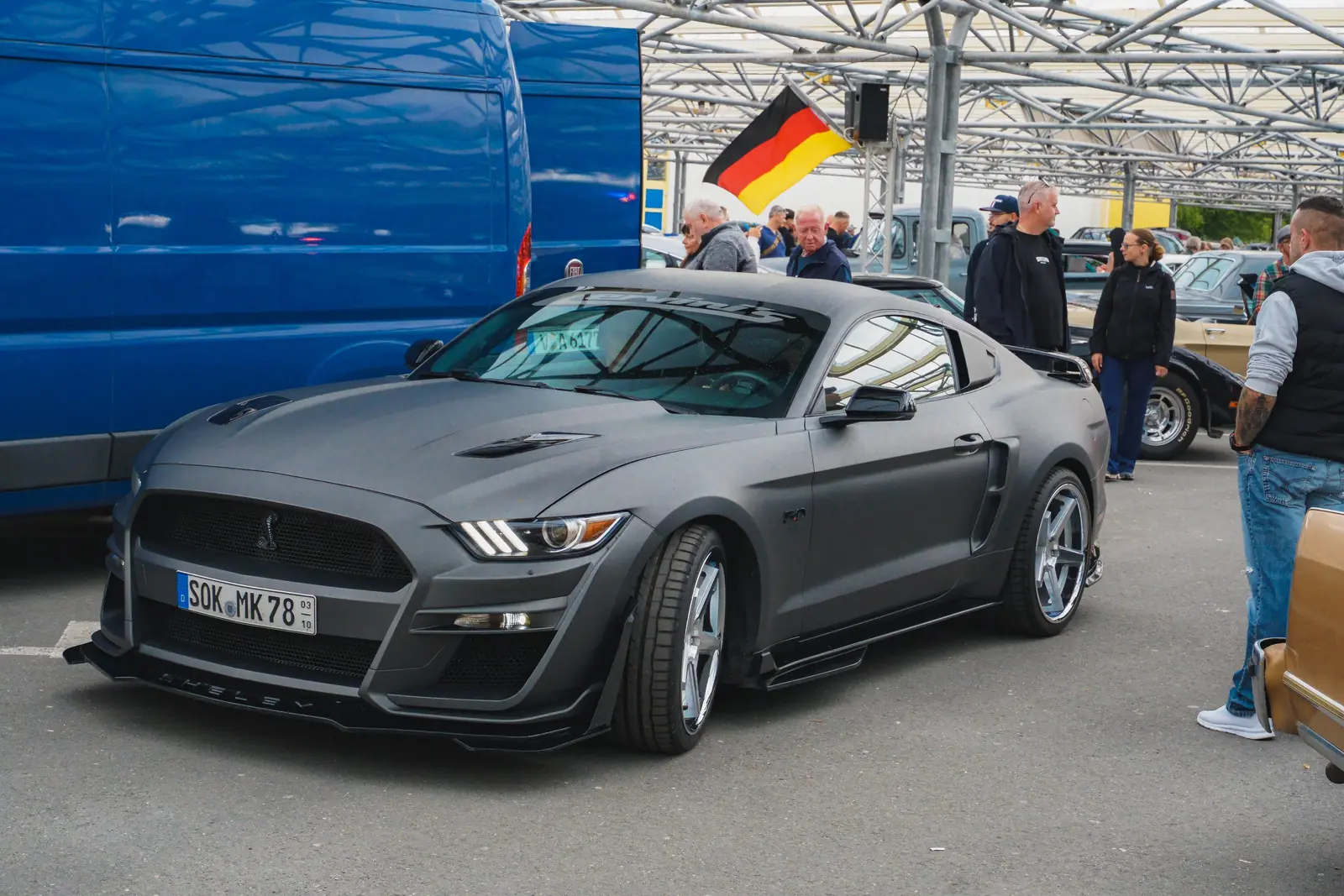 Grauer Mustang im Shelby-Look, dahinter eine Deutschlandfahne