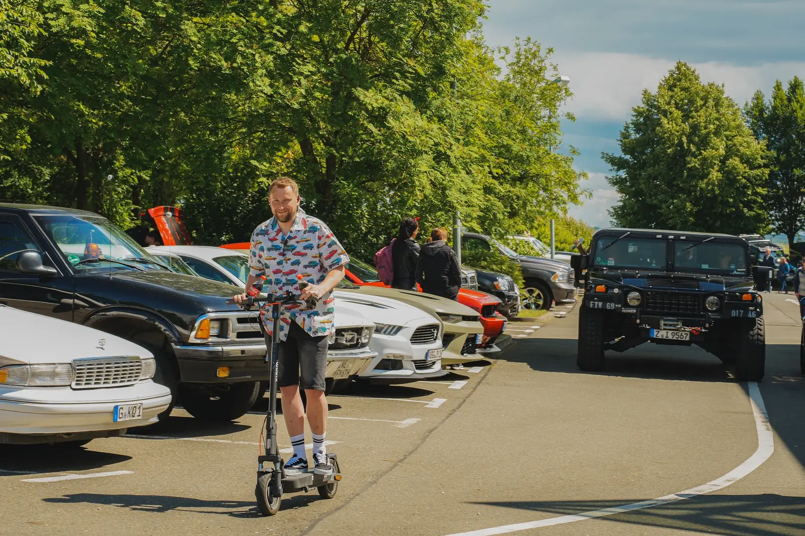 Mann im Hemd mit Automuster auf dem E-Scooter vor geparkten US-Cars
