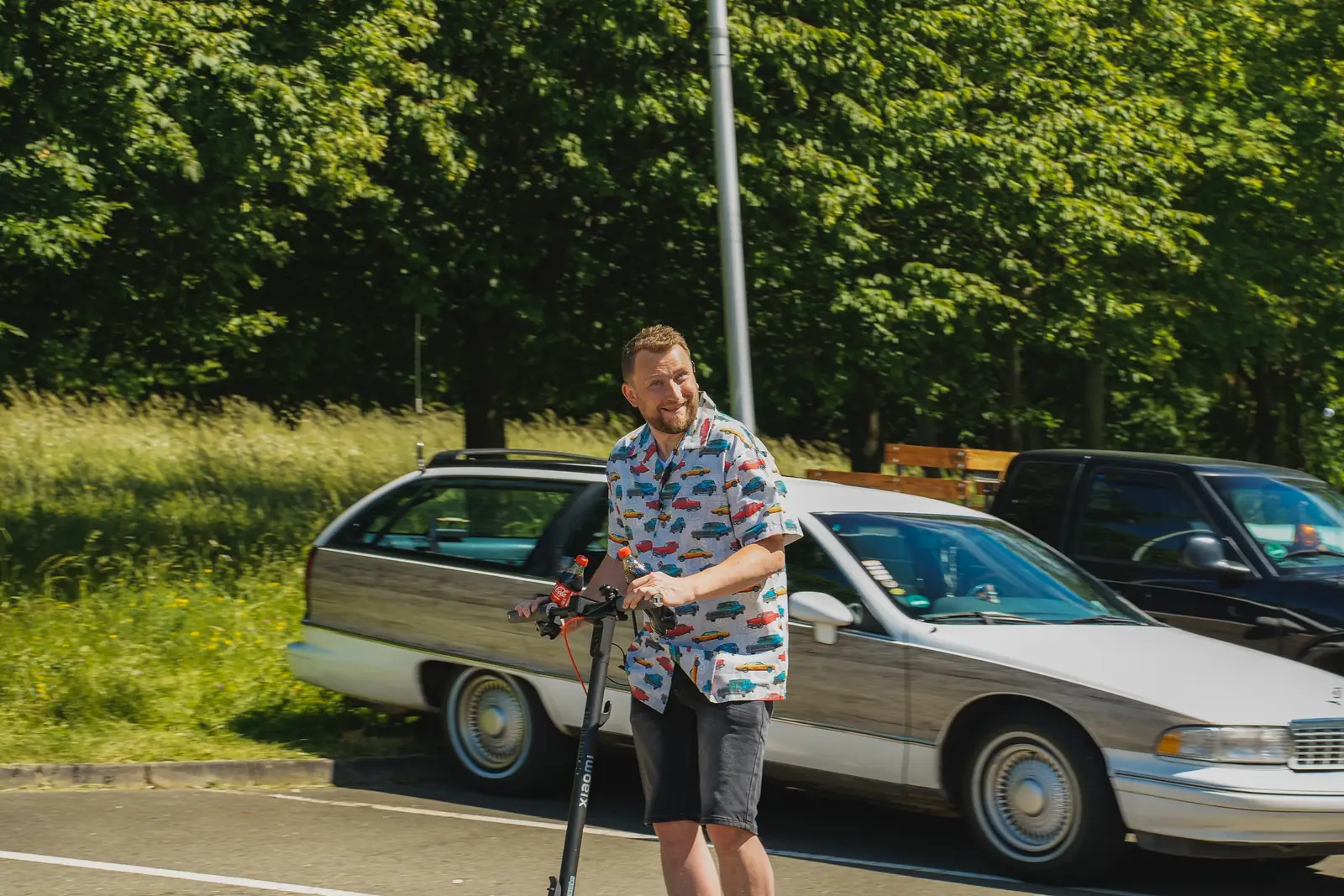 Mann im Hemd mit Automuster auf dem E-Scooter vor einem Chevrolet-Kombi