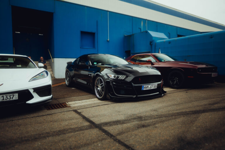 Weiße Corvette und schwarzer Mustang vor blauer Hallenwand